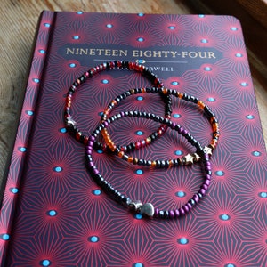 Może przedstawiać: Trzy bransoletki z koralików na książce z czerwoną i czarną okładką. Tytuł książki to "Nineteen Eighty-Four" autorstwa George'a Orwella. Bransoletki są czarne, pomarańczowe i fioletowe.