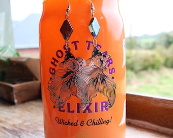 May include: A pair of dangle earrings featuring a fish design. The earrings have a silver-tone finish and are adorned with black and orange fish. The earrings are hanging in front of an orange can with the text "Ghost Tears Elixir Wicked & Chilling!"