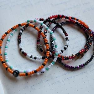 Puede incluir: Un conjunto de cinco pulseras de cuentas de colores con dijes de estrella y ojo. Las pulseras están hechas con cuentas de color naranja, negro, blanco, turquesa y morado.