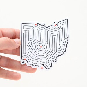 Op de afbeelding: Een witte sticker met een zwarte omtrek van de staat Ohio. De staat is ontworpen als een doolhof met een rode ster in het midden en drie rode stippen langs de rand.