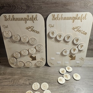 Könnte beinhalten: Zwei Holz-Belohnungstafeln mit den Texten "Belohnungstafel" und den Namen "Luca" und "Luisa". Jede Tafel hat Aussparungen für Holzmarken mit eingravierten Bären und Feen. Zusätzliche Marken mit verschiedenen Tierdesigns liegen darunter.