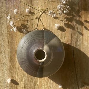 May include: A small, round, ceramic vase with a brown and white glaze. The vase has a wide opening at the top and a narrow opening at the bottom. The vase is sitting on a wooden surface.