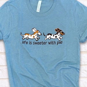 May include: A light blue t-shirt with a graphic of two cartoon dachshunds running. The text "life is sweeter with pie" is printed below the dachshunds.