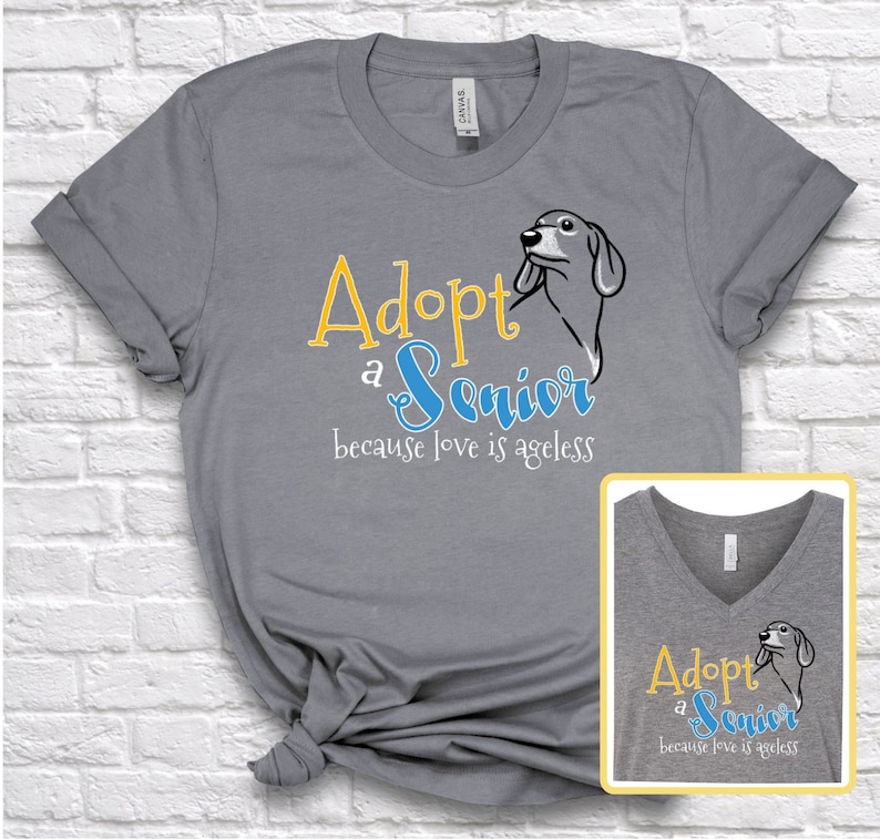 Op de afbeelding: Grijs T-shirt met een blauwe en gele afbeelding met de tekst "Adopt a Senior because love is ageless". De afbeelding bevat ook een cartoonhond.