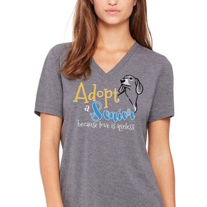 Op de afbeelding: Een grijs V-hals T-shirt met de tekst "Adopt a Senior because love is ageless" in gele en blauwe letters. Naast de tekst staat een cartoon afbeelding van een hond.