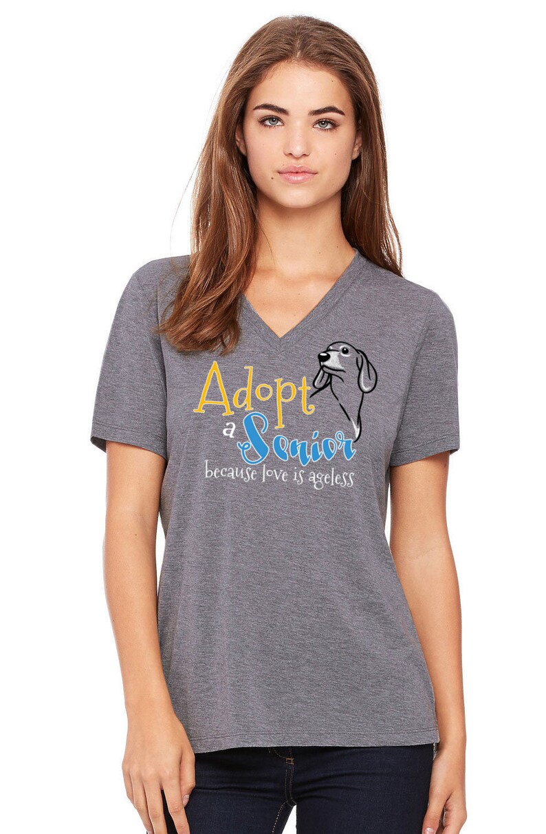 Op de afbeelding: Een grijs V-hals T-shirt met de tekst "Adopt a Senior because love is ageless" in gele en blauwe letters. Naast de tekst staat een cartoon afbeelding van een hond.