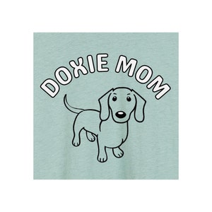 May include: A light blue t-shirt with the text "DOXIE MOM" in white letters with a black outline. A cartoon dachshund is centered below the text.