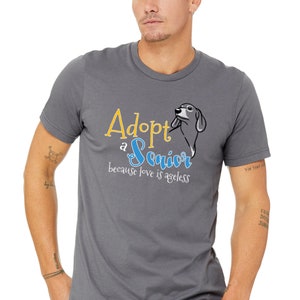 Op de afbeelding: Een grijs T-shirt met de tekst "Adopt a Senior because love is ageless" in gele en blauwe letters. Naast de tekst is een cartoon teckel afgebeeld.
