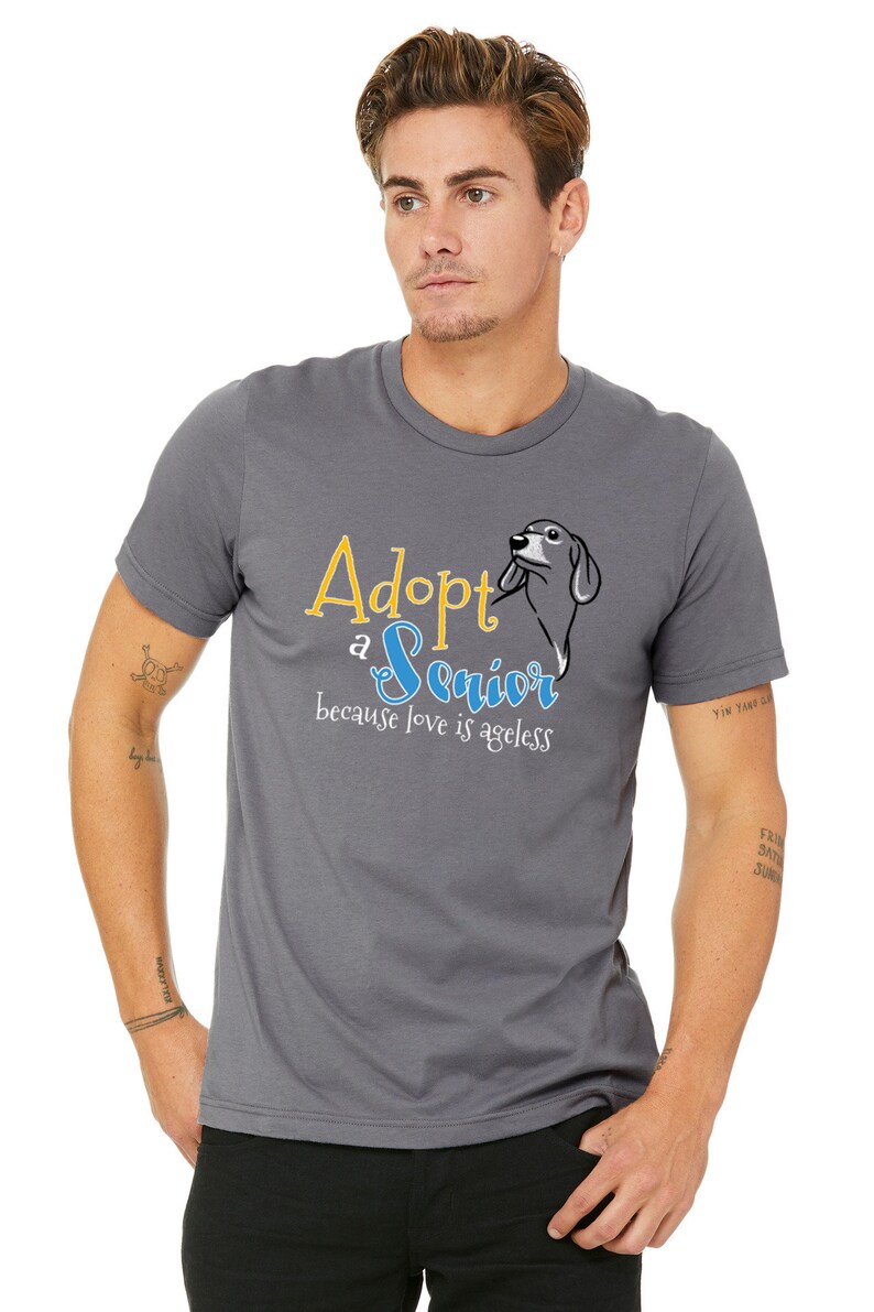 Op de afbeelding: Een grijs T-shirt met de tekst "Adopt a Senior because love is ageless" in gele en blauwe letters. Naast de tekst is een cartoon teckel afgebeeld.