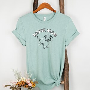 May include: A light green t-shirt with a black and white dachshund illustration and the text "Doxie Mom" in white with a pink outline.