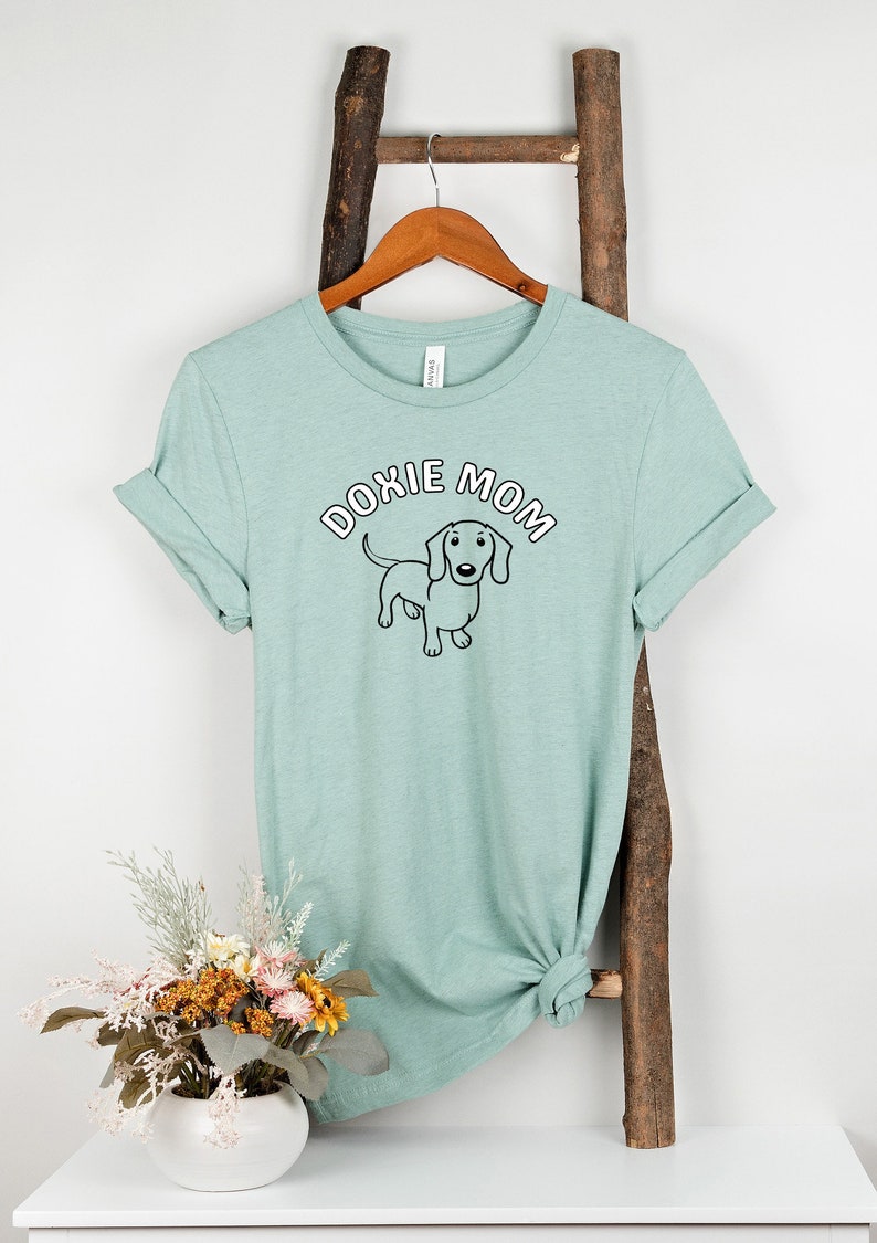 May include: A light green t-shirt with a black and white dachshund illustration and the text "Doxie Mom" in white with a pink outline.