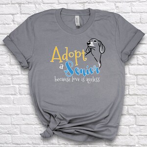 Op de afbeelding: Grijs T-shirt met de tekst "Adopteer een senior, want liefde kent geen leeftijd" in gele en blauwe letters. Rechts van de tekst staat een cartoonafbeelding van een hondenkop.