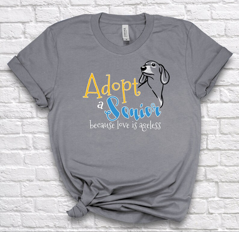 Op de afbeelding: Grijs T-shirt met de tekst "Adopteer een senior, want liefde kent geen leeftijd" in gele en blauwe letters. Rechts van de tekst staat een cartoonafbeelding van een hondenkop.