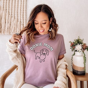 May include: A woman wearing a purple t-shirt with the text "Doxie Mom" and a cartoon dachshund. She is wearing a white cardigan and white pants.
