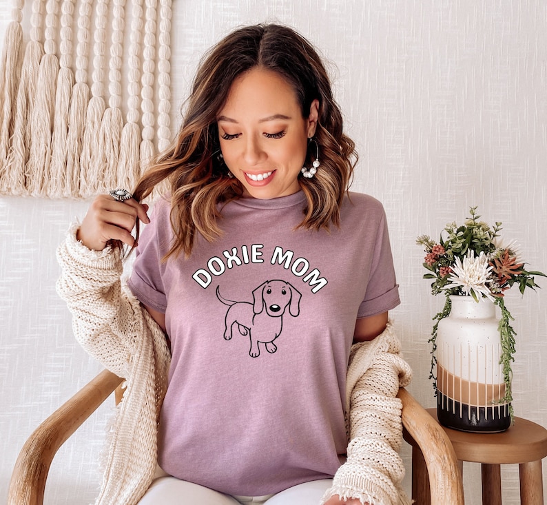 May include: A woman wearing a purple t-shirt with the text "Doxie Mom" and a cartoon dachshund. She is wearing a white cardigan and white pants.