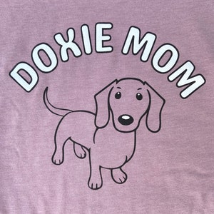 May include: A purple t-shirt with the text "Doxie Mom" in white letters and a cartoon dachshund in black outline.