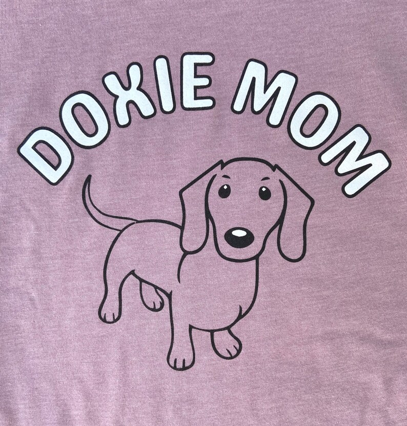 May include: A purple t-shirt with the text "Doxie Mom" in white letters and a cartoon dachshund in black outline.