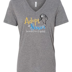 Op de afbeelding: Grijs V-hals T-shirt met de tekst "Adopteer een senior, want liefde kent geen leeftijd" in gele en blauwe letters. Een cartoon teckel is te zien in het ontwerp.