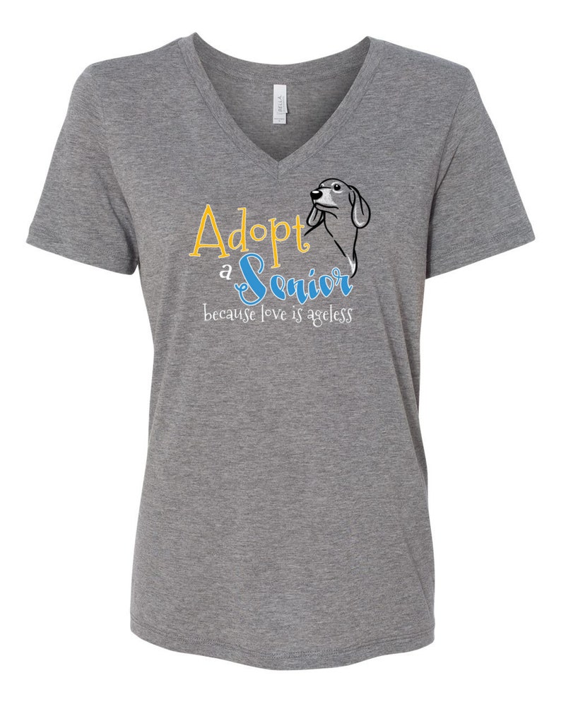 Op de afbeelding: Grijs V-hals T-shirt met de tekst "Adopteer een senior, want liefde kent geen leeftijd" in gele en blauwe letters. Een cartoon teckel is te zien in het ontwerp.
