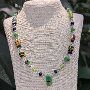 May include: A colourful beaded necklace featuring green, yellow, black, and red beads. The necklace has a silver chain and a green pendant. The necklace is displayed on a mannequin.