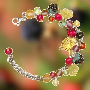 Puede incluir: Una pulsera de cadena en tono plateado con un surtido de dijes de vidrio de colores, incluyendo hojas amarillas translúcidas, bayas rojas, moras oscuras y varias cuentas esféricas en tonos amarillos, rojos y verdes con un cierre de mosquetón.