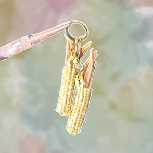 May include: A pair of handmade earrings featuring miniature corn on the cob designs. The earrings have a gold-colored hoop and a charm. The corn is made of yellow and white beads, with brown husk details. The earrings are held by a tool.