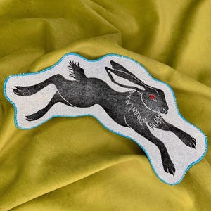 Peut inclure: Écusson noir et blanc représentant un lièvre courant avec un œil rouge. Le patch a une bordure bleu clair et est sur fond blanc. Le lièvre est en plein saut.