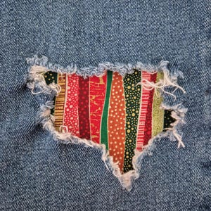 May include: Close-up of a denim fabric with a distressed patch. The patch is made of colorful fabric strips in red, green, and gold, with various patterns. The edges of the denim are frayed, adding a vintage look.