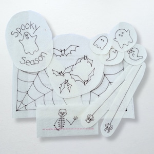 May include: A selection of white fabric patches featuring black embroidered Halloween designs. Includes ghosts, bats, a spider web, a skeleton, spiders, and the text "Spooky Season". Suitable for crafting or seasonal decorations.