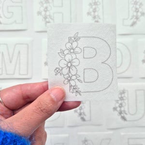 May include: A white fabric square with a grey outline of the letter B decorated with a floral design. The flowers are stylised and have a simple design.