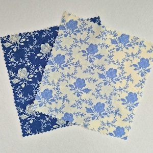 May include: Two fabric swatches with floral patterns. One swatch is navy blue with white roses and vines. The other is cream-colored with blue roses and vines. Both have a scalloped edge.