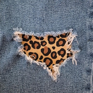 May include: Close-up of a distressed denim fabric with a leopard print patch. The blue denim has a torn section revealing a tan fabric with brown spots. The edges of the tear are frayed, adding texture to the design.