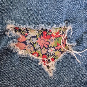 May include: Close-up of a denim fabric with a distressed area patched with floral fabric. The patch features a pattern of pink, red, and blue flowers on a dark blue background. White threads are visible.