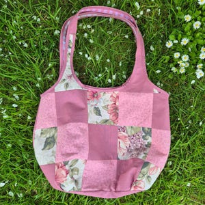 May include: A patchwork tote bag featuring a combination of solid dusty rose fabric squares and floral patterned fabric squares in shades of pink, green, and white. The bag has two shoulder straps with a pink and white striped interior lining.