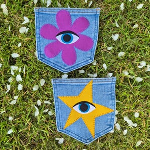 May include: Two blue denim pockets featuring fabric appliqués. The top pocket displays a purple flower with a central eye motif. The bottom pocket features a yellow star with a central eye motif. Both pockets have visible stitching along the edges.