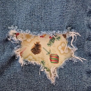 May include: Close-up of a distressed denim patch with a Christmas-themed fabric insert. The fabric features pinecones, ornaments, and sprigs on a cream background. The denim is a medium blue with frayed edges.