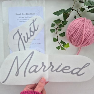 May include: Two white fabric patches with the words "Just" and "Married" in gray script. A pink ball of yarn and green leaves are in the background. The patches are from Beech Tree Handmade.