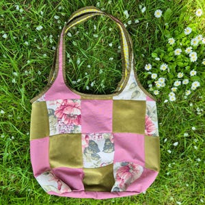 May include: A patchwork tote bag featuring a grid of alternating solid olive green squares and floral patterned squares in shades of pink, white, and green. The bag has two matching fabric handles with a yellow and white polka dot interior lining.