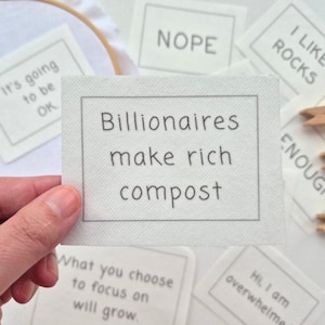 May include: Embroidered white fabric squares with text. One says "Billionaires make rich compost", another "Nope", and others read "It's going to be OK" and "I like rocks".
