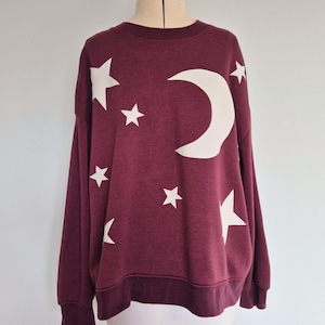 May include: Burgundy sweatshirt with a white crescent moon and star design. The long-sleeved top has a crew neck and ribbed cuffs and hem. The celestial-themed graphic is printed on the front of the garment.