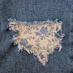 May include: Close-up of distressed blue denim fabric with a floral patch. The patch is a light cream color with a blue floral pattern. The edges of the denim are frayed, adding a vintage aesthetic.
