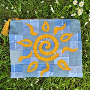 May include: A rectangular zipped pouch made of patchwork denim squares in various shades of blue. The front features a large, embroidered yellow sun design with a central spiral and radiating flame-like rays. A yellow tassel is attached to the zip.