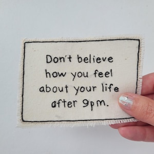 May include: An off-white embroidered patch with black stitching, rectangular in shape. The patch displays the text: "Don't believe how you feel about your life after 9pm." The edges are frayed. The patch is held by a hand.