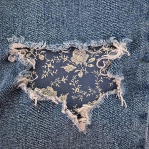 May include: A section of blue denim fabric featuring a distressed, frayed hole. Visible through the opening is a dark blue fabric patch decorated with a light-colored, intricate floral pattern consisting of roses and winding leafy vines.