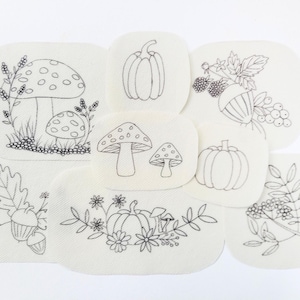 May include: A selection of embroidered patches with autumnal designs. The patches display detailed black line drawings of mushrooms, pumpkins, acorns, berries, and foliage on a cream fabric. The patches are in various shapes.