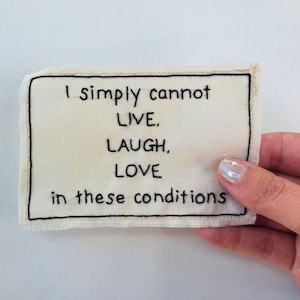 May include: A rectangular, off-white fabric patch with black embroidered text that reads "I simply cannot LIVE, LAUGH, LOVE in these conditions." The patch has a black border and a raw edge.