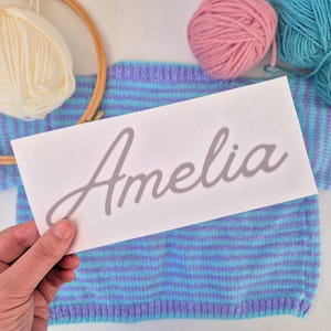 May include: A white label with the name "Amelia" in grey script is held over a blue and purple striped knitted garment. Balls of pink, blue, and cream yarn are visible, along with an embroidery hoop.
