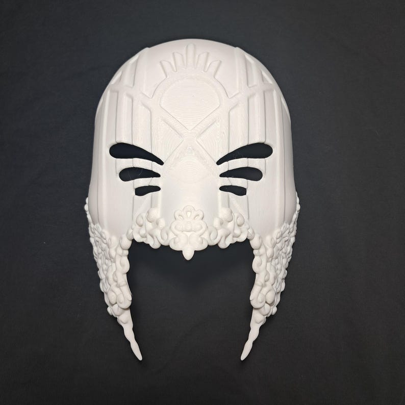 May include: White plastic mask with intricate details and a geometric design. The mask has eye holes and a pointed chin.