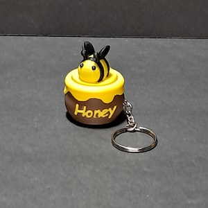 May include: A honey pot keychain with a cartoon bee on top. The honey pot is brown with yellow accents and the word "Honey" in yellow. The bee is yellow and black. A silver key ring is attached.