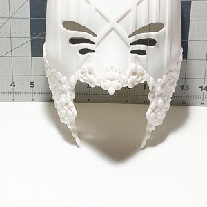 May include: White plastic mask with a detailed design. The mask has two eye holes and a decorative border around the bottom.